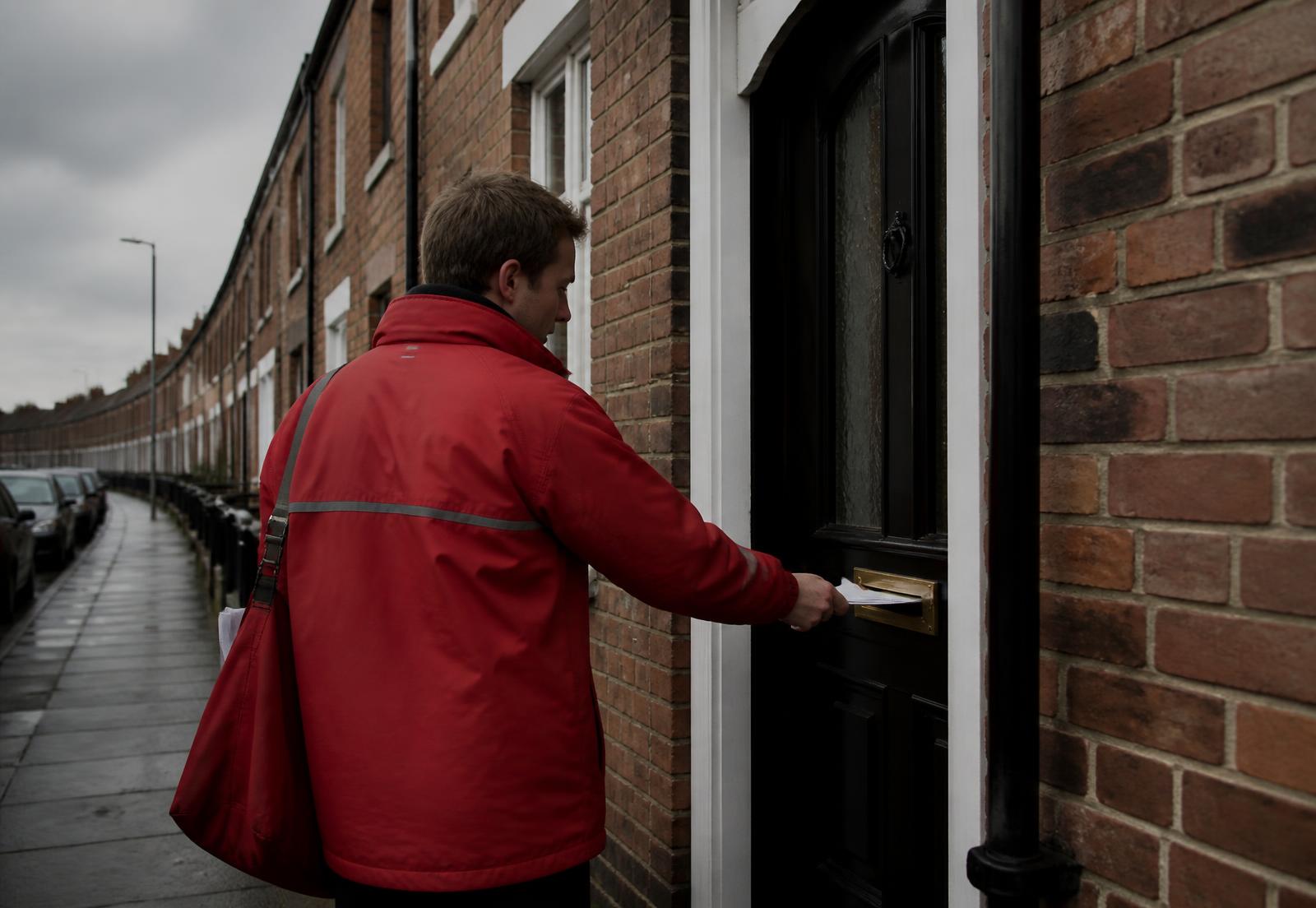 Delivery worker in a red raincoat posting printed mail through the letterbox of a UK terraced house on an overcast day